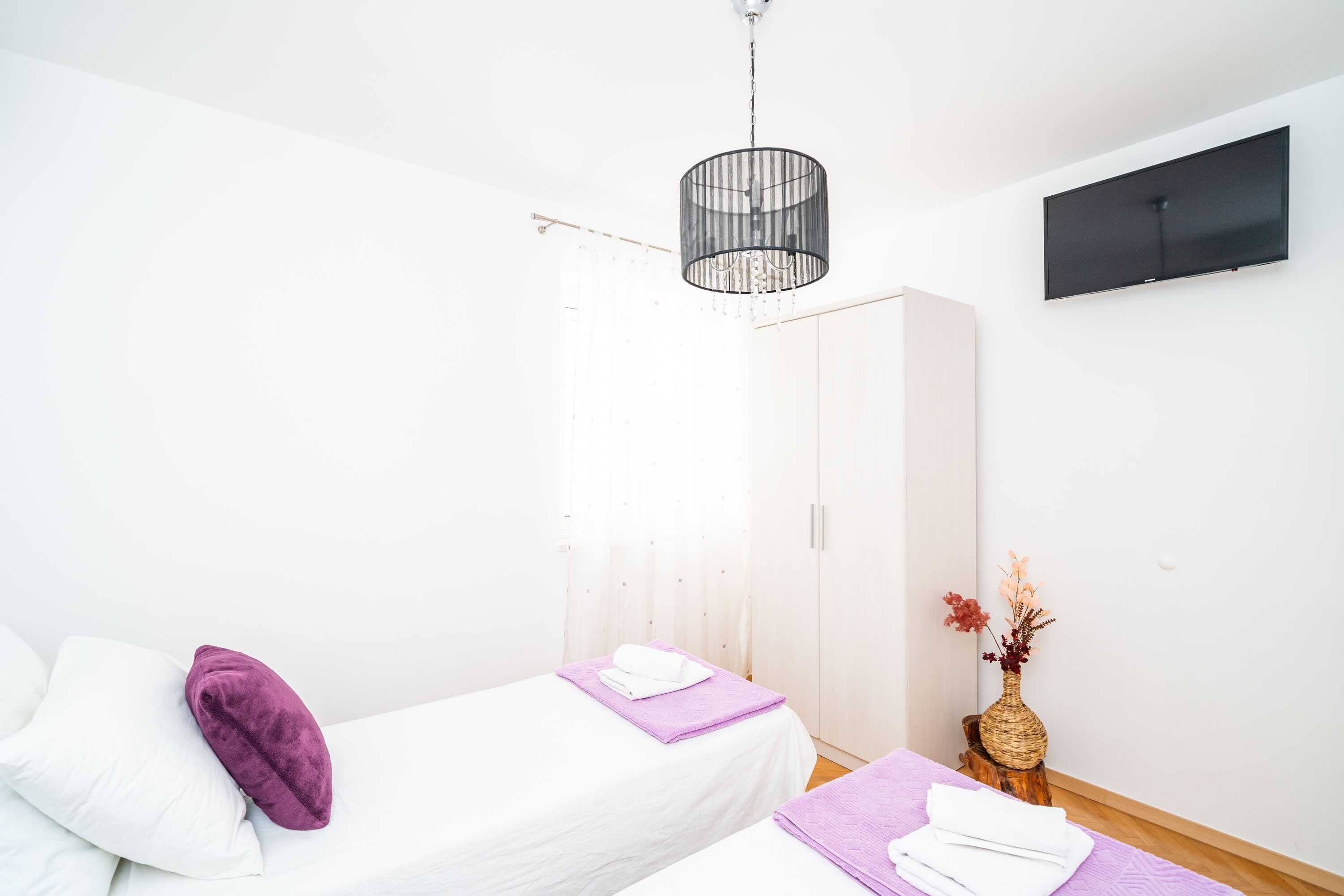 A modern bedroom with two beds, a TV, a hanging light fixture, and a wooden stool with a vase.