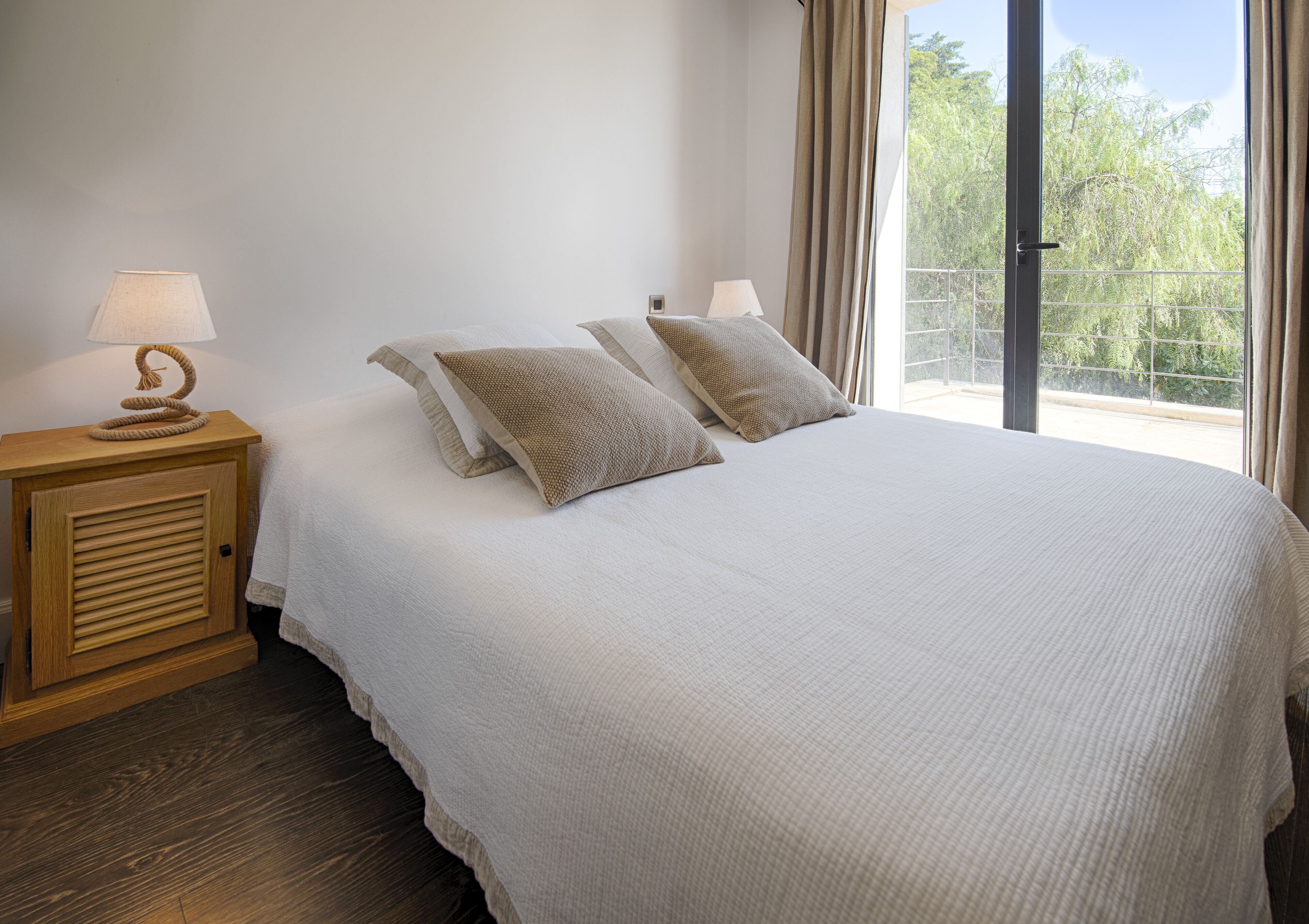 A bedroom with a large bed, two bedside lamps, a wooden nightstand, and a view of greenery through a balcony door.