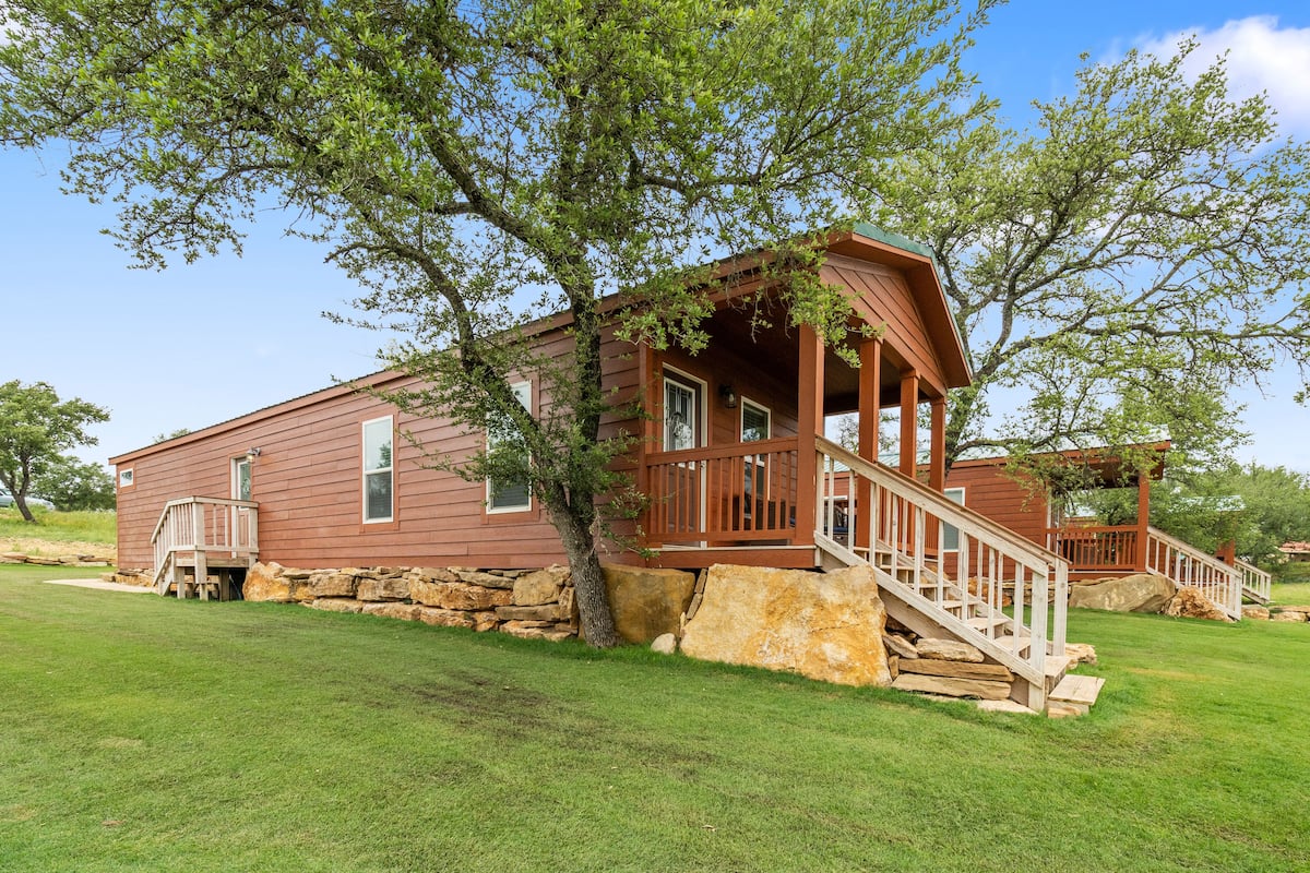 A wooden cabin with a porch and steps, surrounded by a stone wall and a large tree.