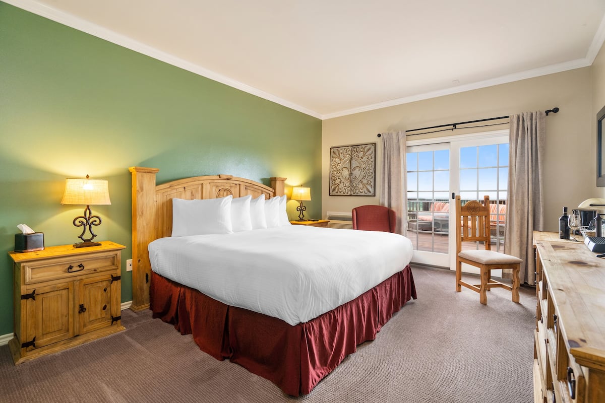 A hotel room with a large bed, wooden headboard, two bedside lamps, a wooden nightstand, a chair, a desk with a lamp, and a window with curtains.