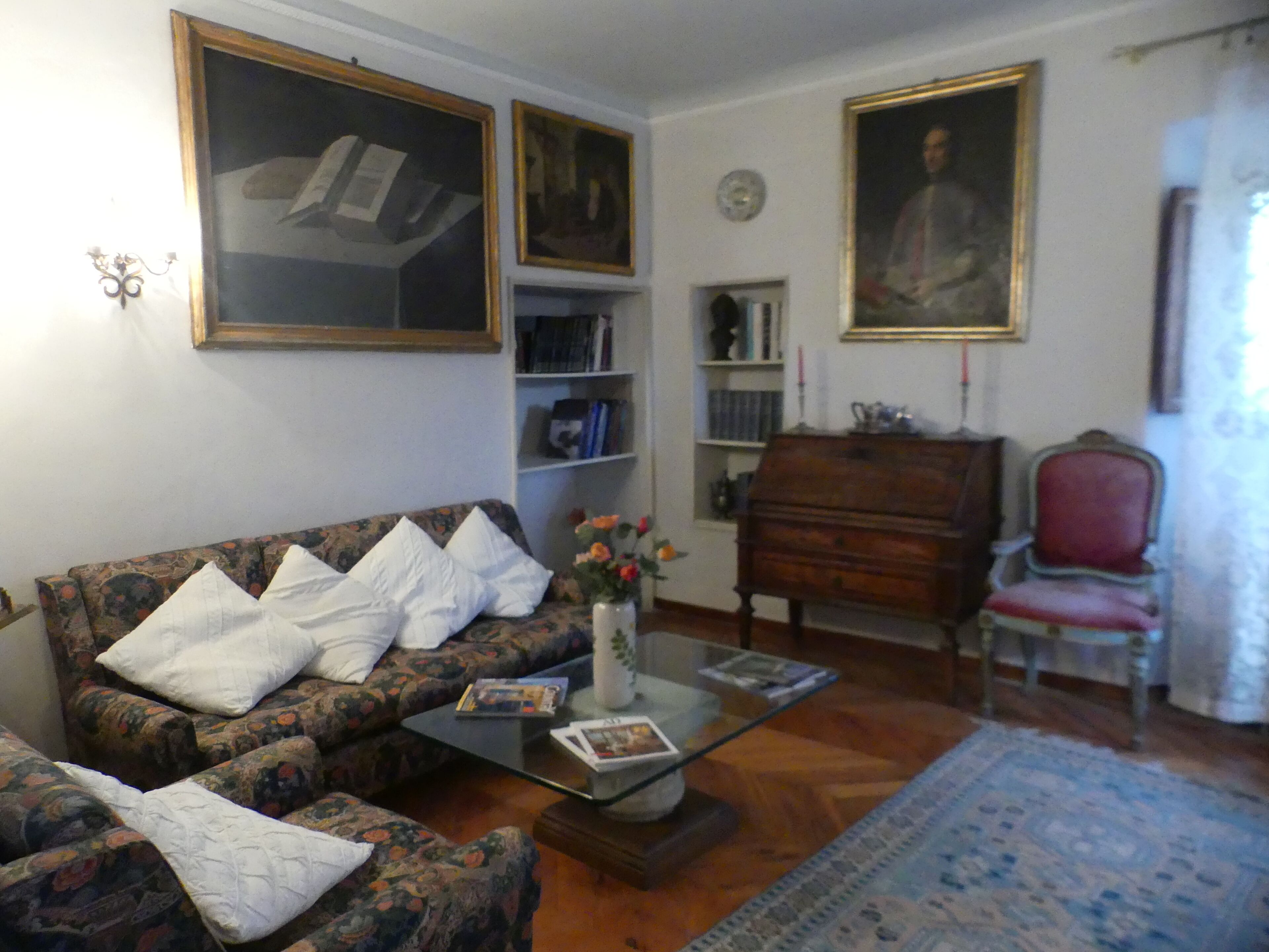 Un salon avec un canapé à motifs floraux, une table basse en verre, un fauteuil rouge, un meuble en bois et un grand portrait encadré accroché au mur.