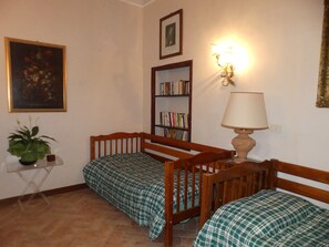 Una habitación con dos camas de madera, una estantería, un cuadro enmarcado, una mesa con una planta y una lámpara de pared.