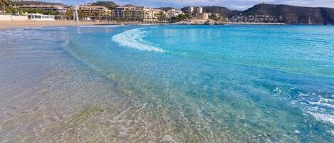 Une plage avec une eau turquoise et limpide, un rivage sablonneux et des bâtiments en arrière-plan.