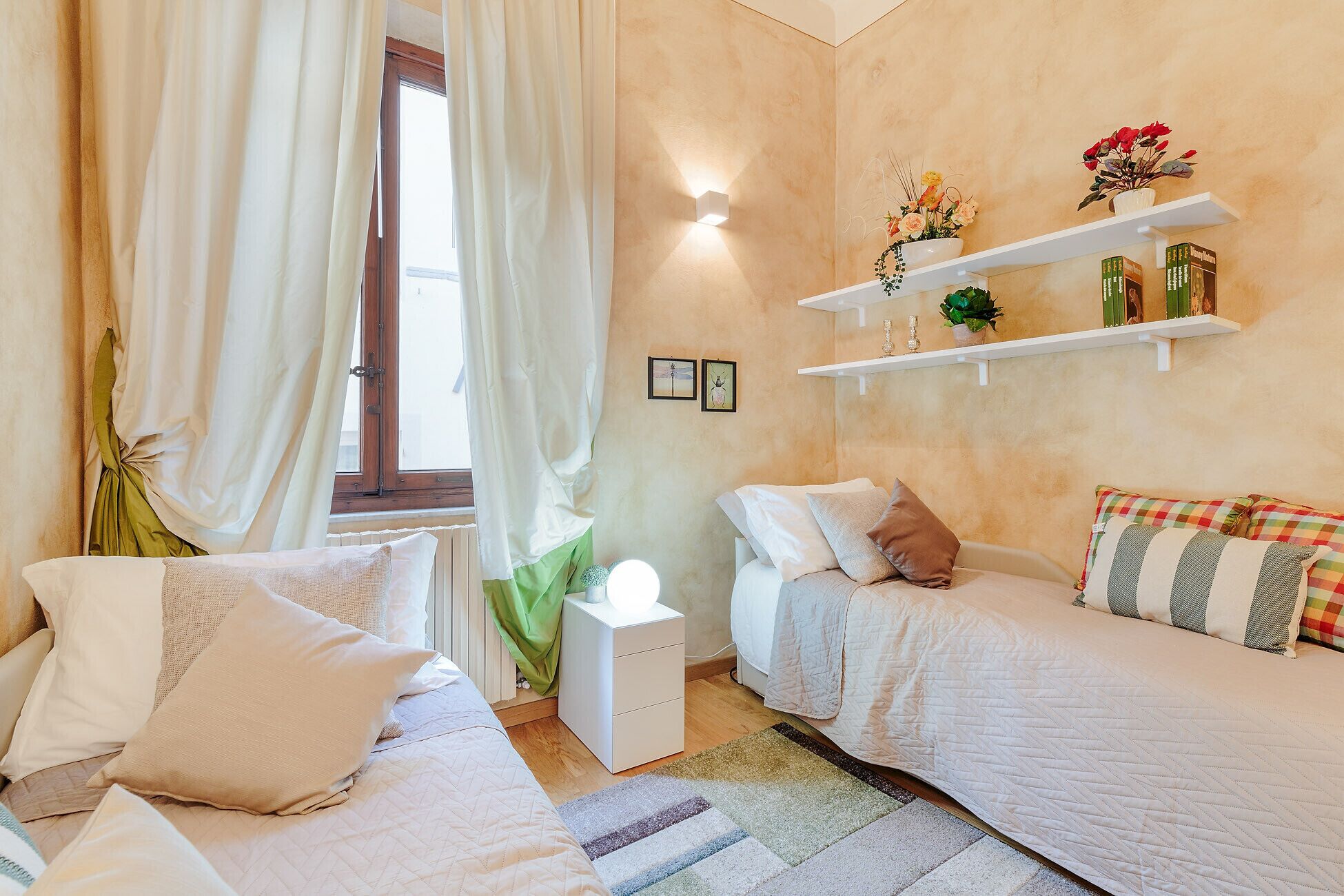 Ein Schlafzimmer mit zwei Betten, einem Nachttisch, Vorhängen an einem Fenster, an der Wand befestigten Regalen mit Pflanzen und einer Wandleuchte.