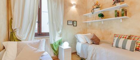 Ein Schlafzimmer mit zwei Betten, einem Nachttisch, Vorhängen an einem Fenster, an der Wand befestigten Regalen mit Pflanzen und einer Wandleuchte.