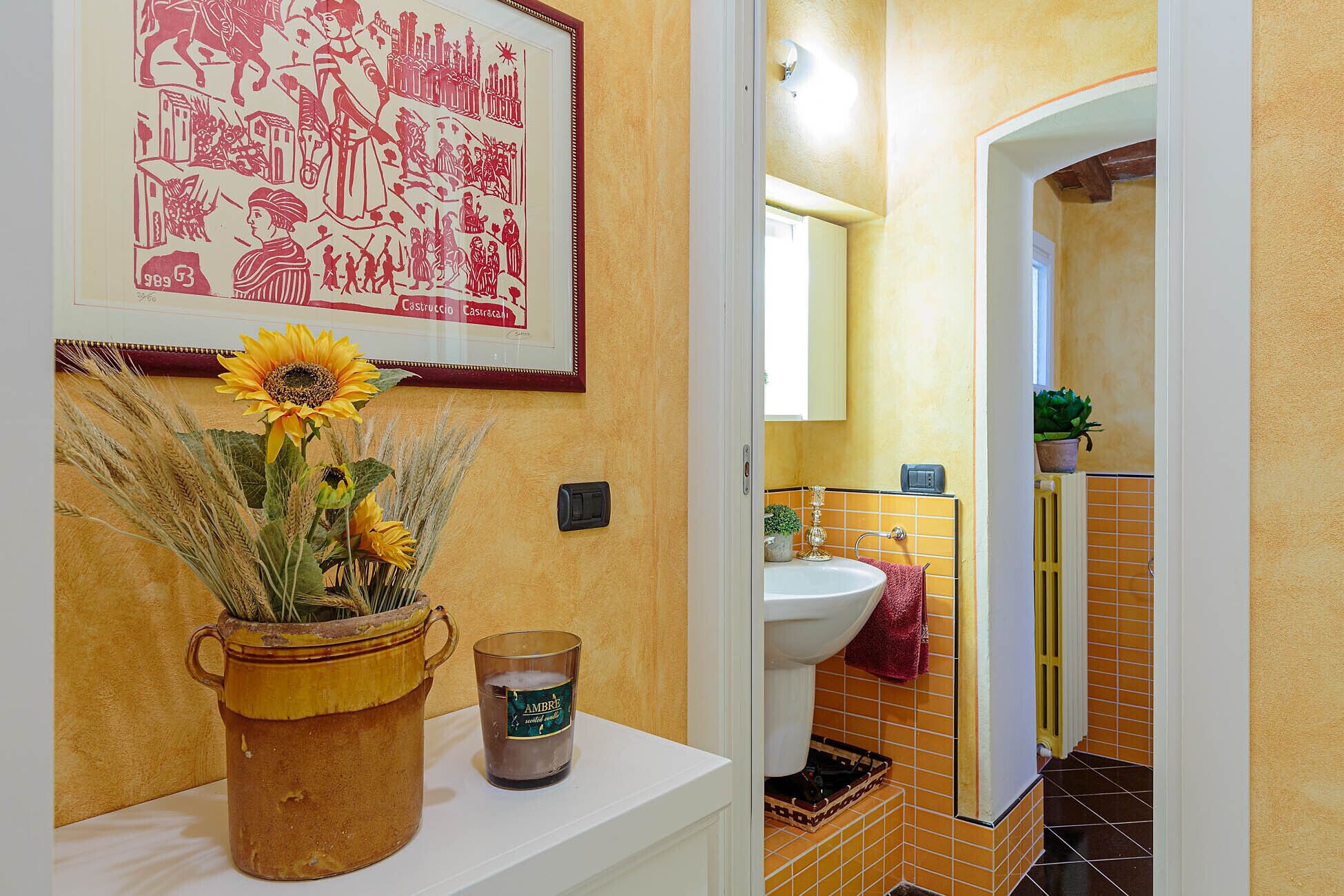 A bathroom with a yellow tiled wall, a sink, and a mirror. There is a framed artwork above the sink and a vase with sunflowers on the counter.