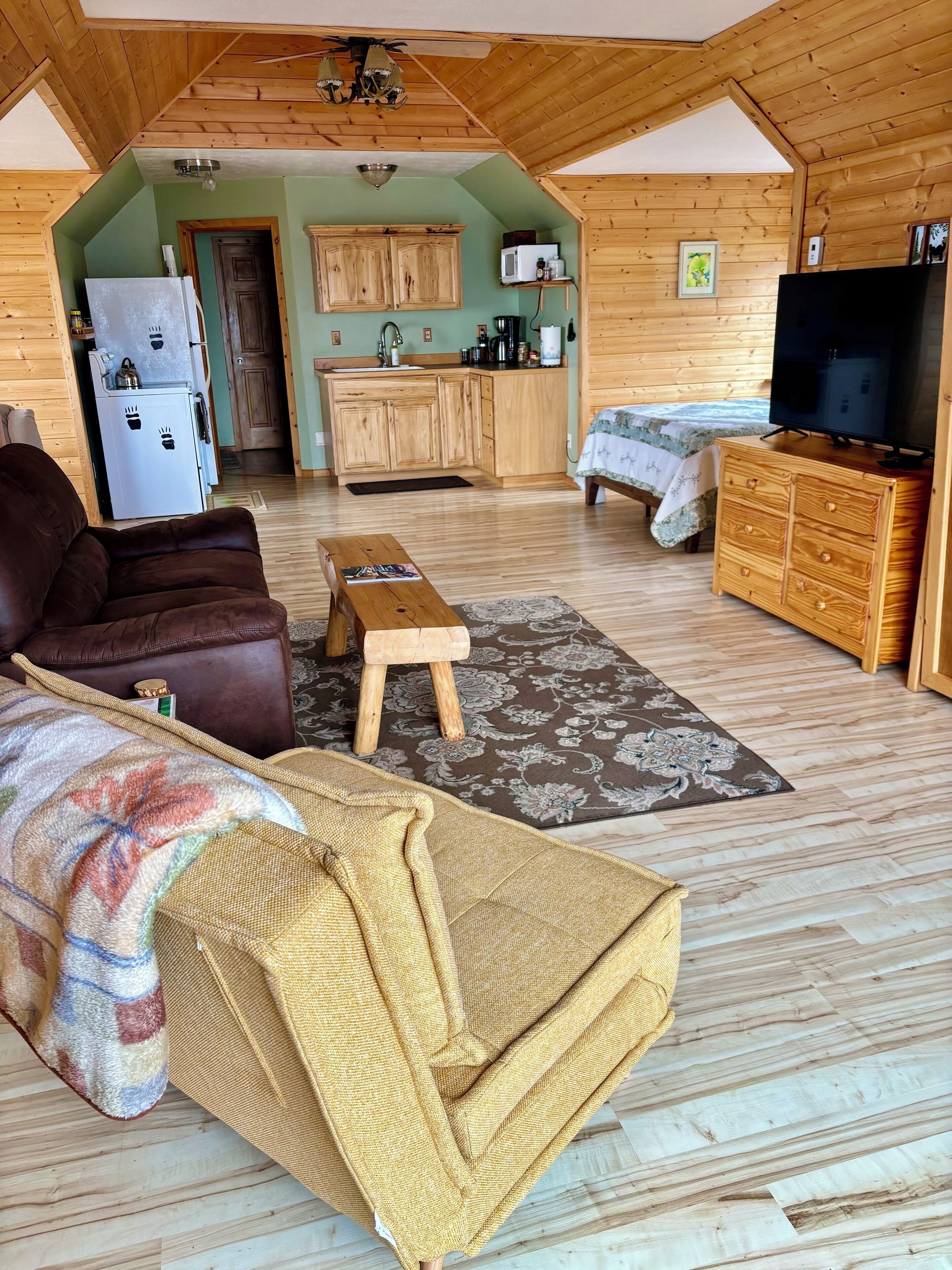 A cozy living room with a brown leather sofa, a wooden coffee table, and a patterned rug. A television is on a wooden cabinet, and a kitchen area is visible in the background.