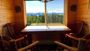 A cabin with a large window offering a view of a forest and mountains. There are two wooden chairs and a table in the center.