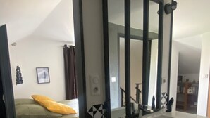Ein modernes Badezimmer mit einem großen Spiegel, einem Waschbecken und einer Toilette. Im angrenzenden Raum befindet sich ein Bett mit grüner Bettwäsche und einem gelben Kopfkissen.