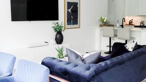 Ett modernt vardagsrum med en mörkblå soffa, en ljusblå fåtölj, en platt-TV och ett minimalistiskt köksområde.