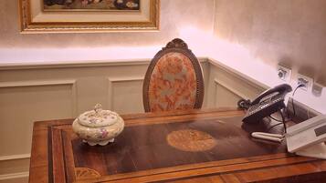 A wooden desk with a telephone, a decorative bowl, and a chair with floral upholstery. A framed painting is on the wall above the desk.