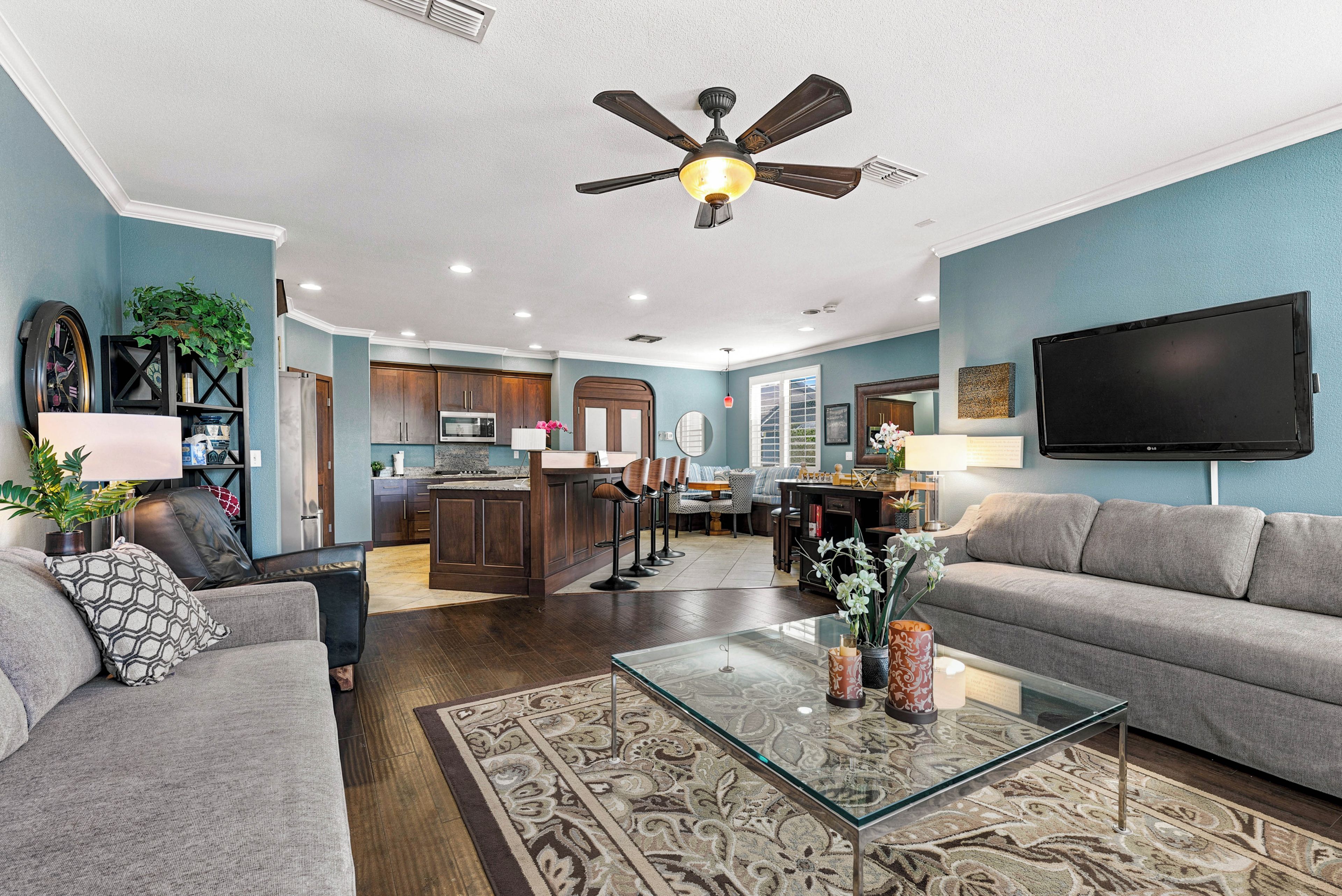 A modern living room with a glass coffee table, a ceiling fan, and a kitchen in the background.