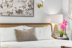 Un lit bien fait, avec une tête de lit, une lampe fixée au mur et un tableau floral au-dessus.