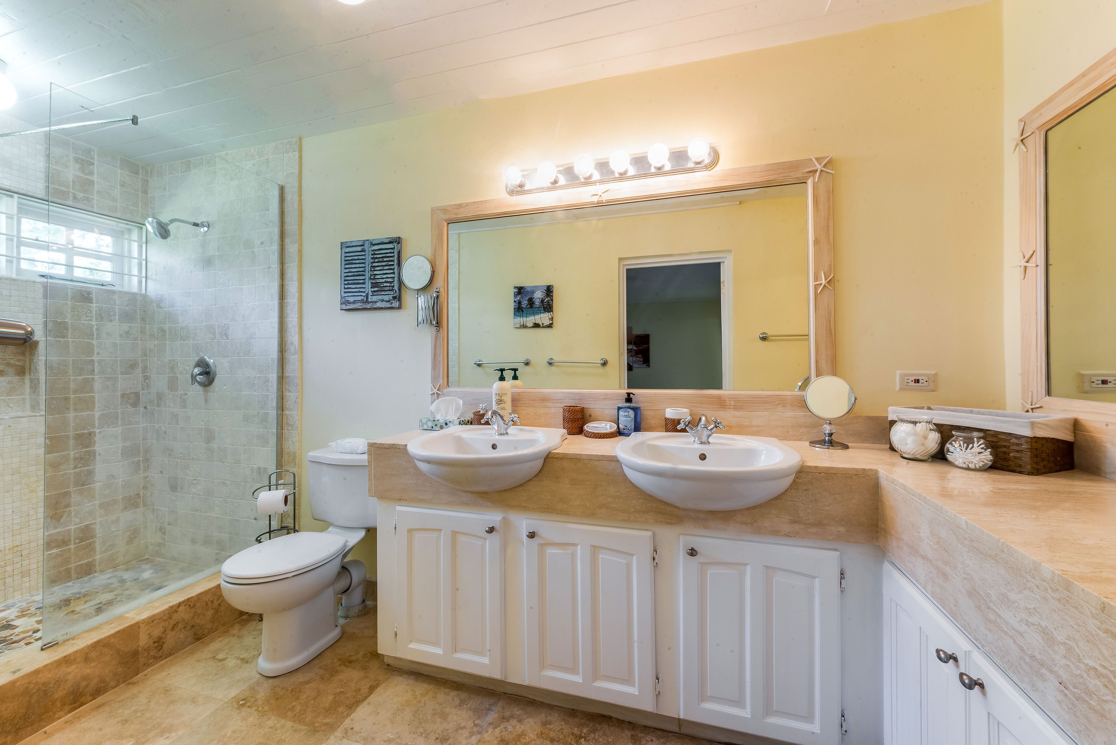 Un baño con cabina de ducha de vidrio, dos lavamanos, un inodoro y un espejo grande con luces superiores.