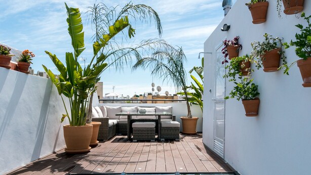Una terraza en la azotea con barandilla de vidrio, plantas en macetas, mobiliario exterior y cielo despejado.