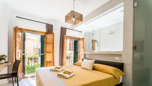 Un dormitorio moderno con una cama grande, un escritorio y una silla. Cuenta con un balcón con contraventanas verdes y vistas a edificios.