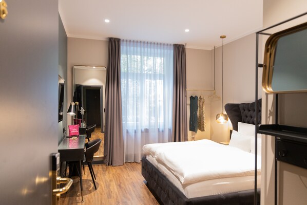 A hotel room with a bed, a desk with a chair, a mirror, a wardrobe, and a window with curtains. - Munich Rooms Hotel (Munich)