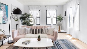 A modern living room with a light pink sofa, a white coffee table, and a patterned rug. There are two large windows with sheer curtains, a floor lamp, and a potted plant. - Sun filled + Airy Large 2 Bedroom apt in West adam’s (Los Angeles)