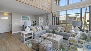A spacious living room with a large gray sectional sofa, a white coffee table, and a view of palm trees through large windows.