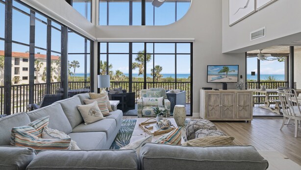 A spacious living room with a large window overlooking a beach, a ceiling fan, and a view of the ocean.