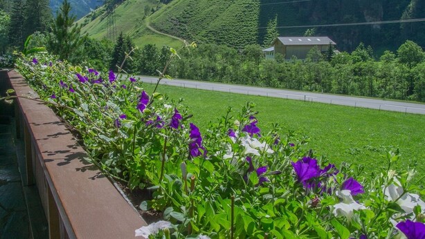 A balcony with a wooden railing overlooking a mountain landscape with a road and a building. - 110m² vacation apartment in a quiet location in the wild and romantic Zillertal valley (Mayrhofen)