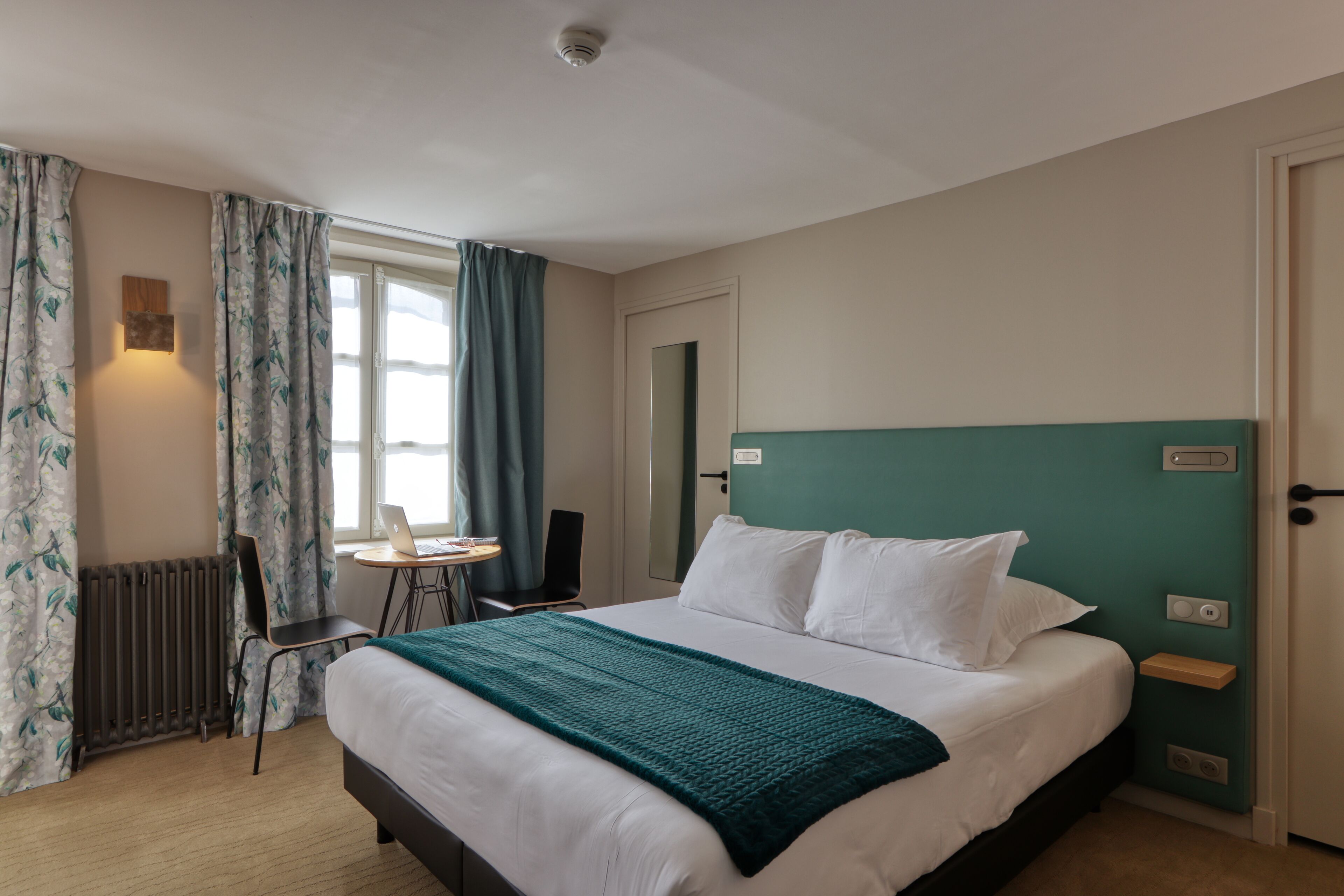 Habitación de hotel con cama, escritorio con una computadora portátil, silla, radiador y ventana con cortinas.