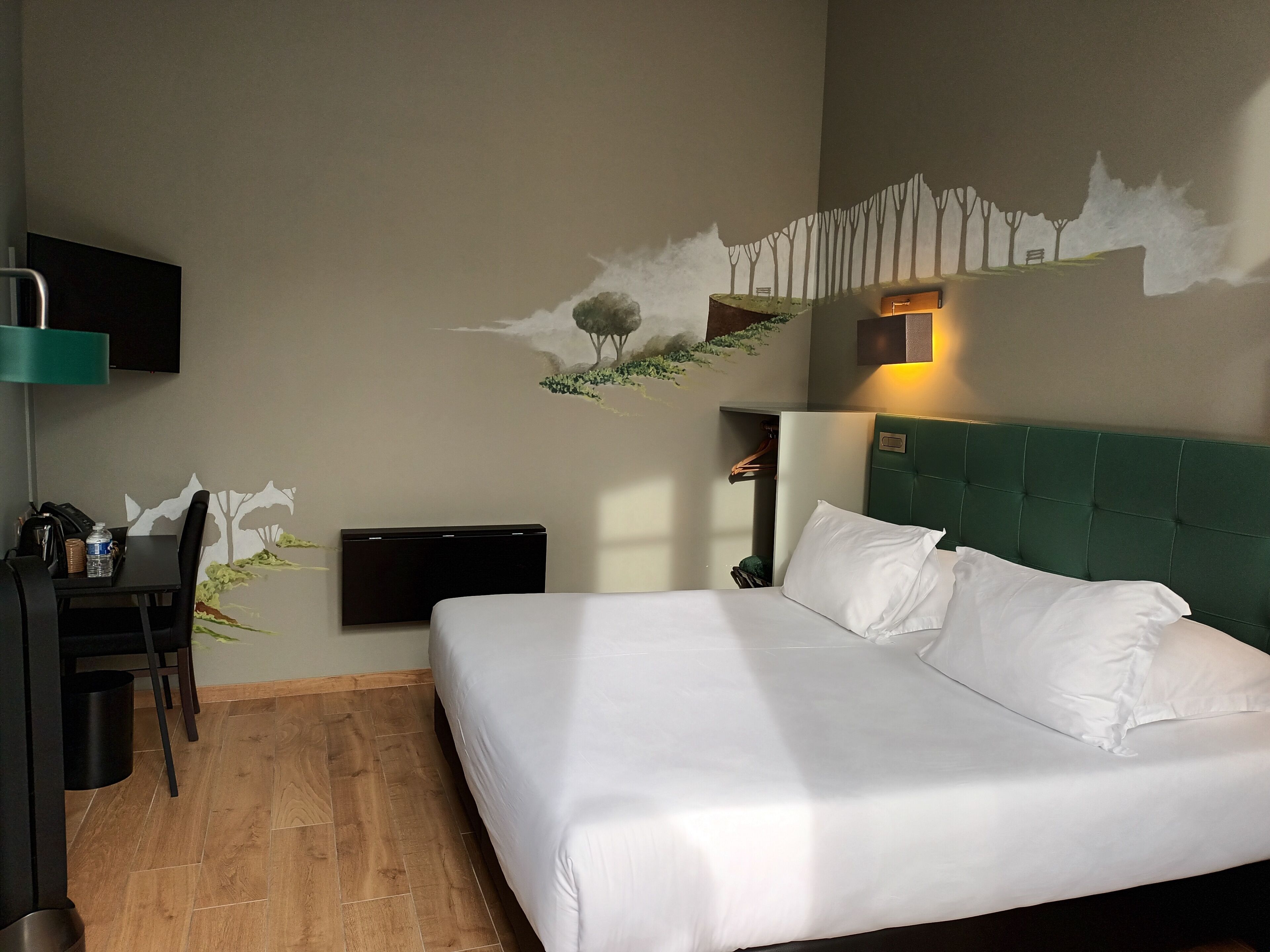 A hotel room with a bed, a desk, a chair, a TV, and a wall-mounted shelf.