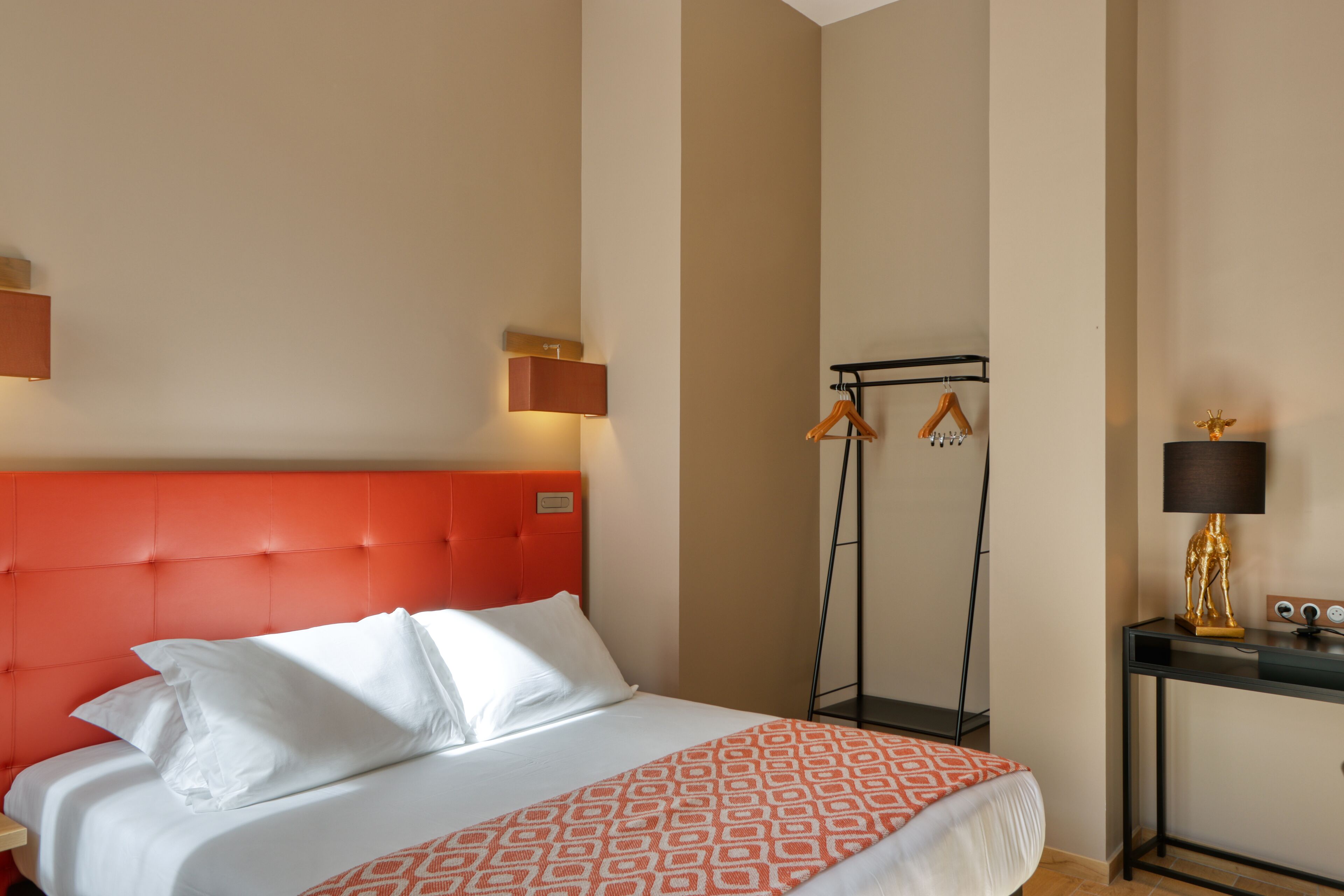A hotel room with a red upholstered headboard, a bed with white linens, a patterned throw, a bedside table with a lamp, and a coat rack with hangers.