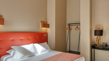 Una habitación de hotel con un cabecero tapizado en rojo, una cama con ropa de cama blanca, una manta estampada, una mesita de noche con lámpara y un perchero con ganchos.