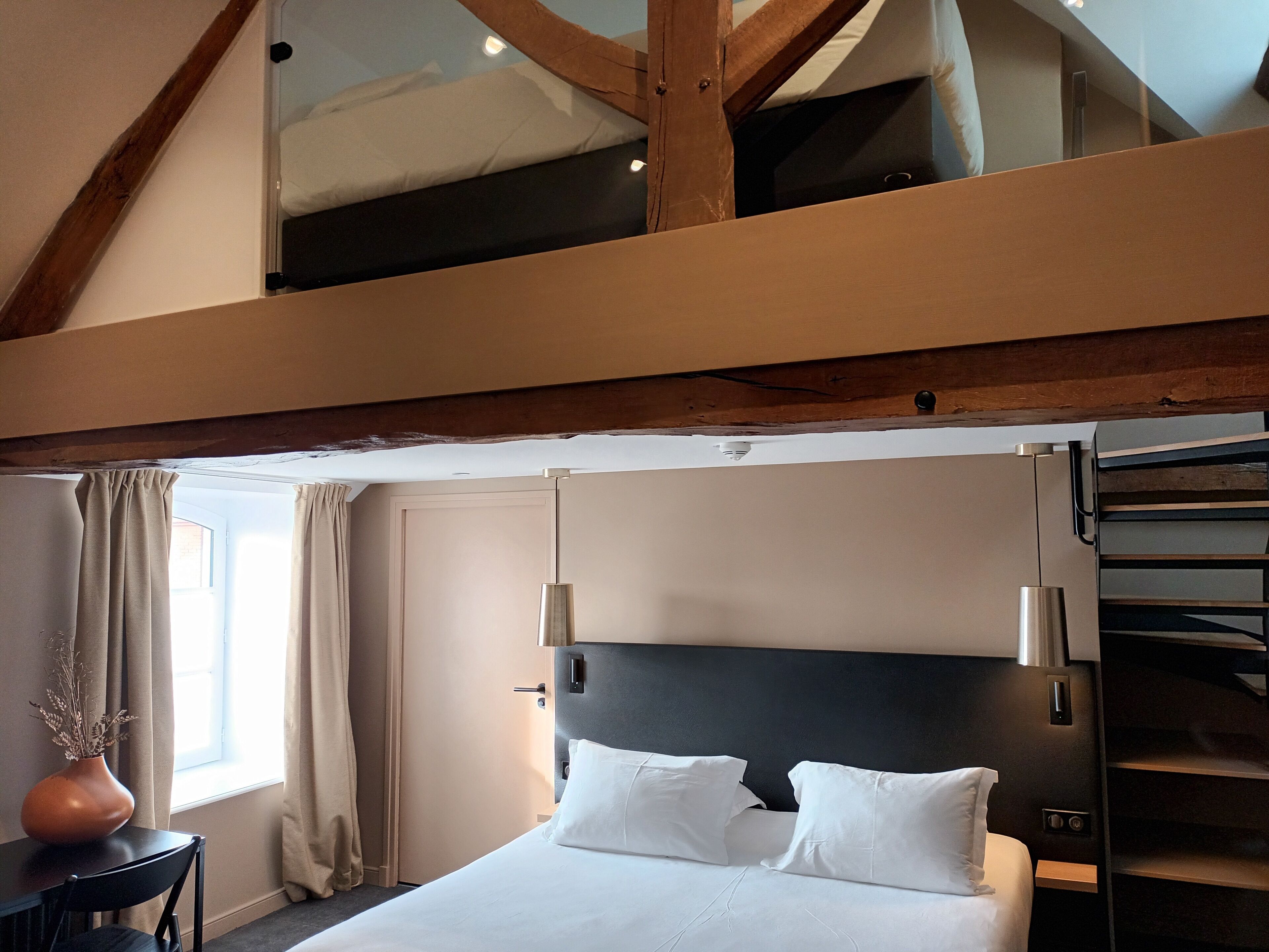 Quarto de hotel moderno com uma cama grande, uma escrivaninha, um vaso com plantas secas e vista do céu pela janela.