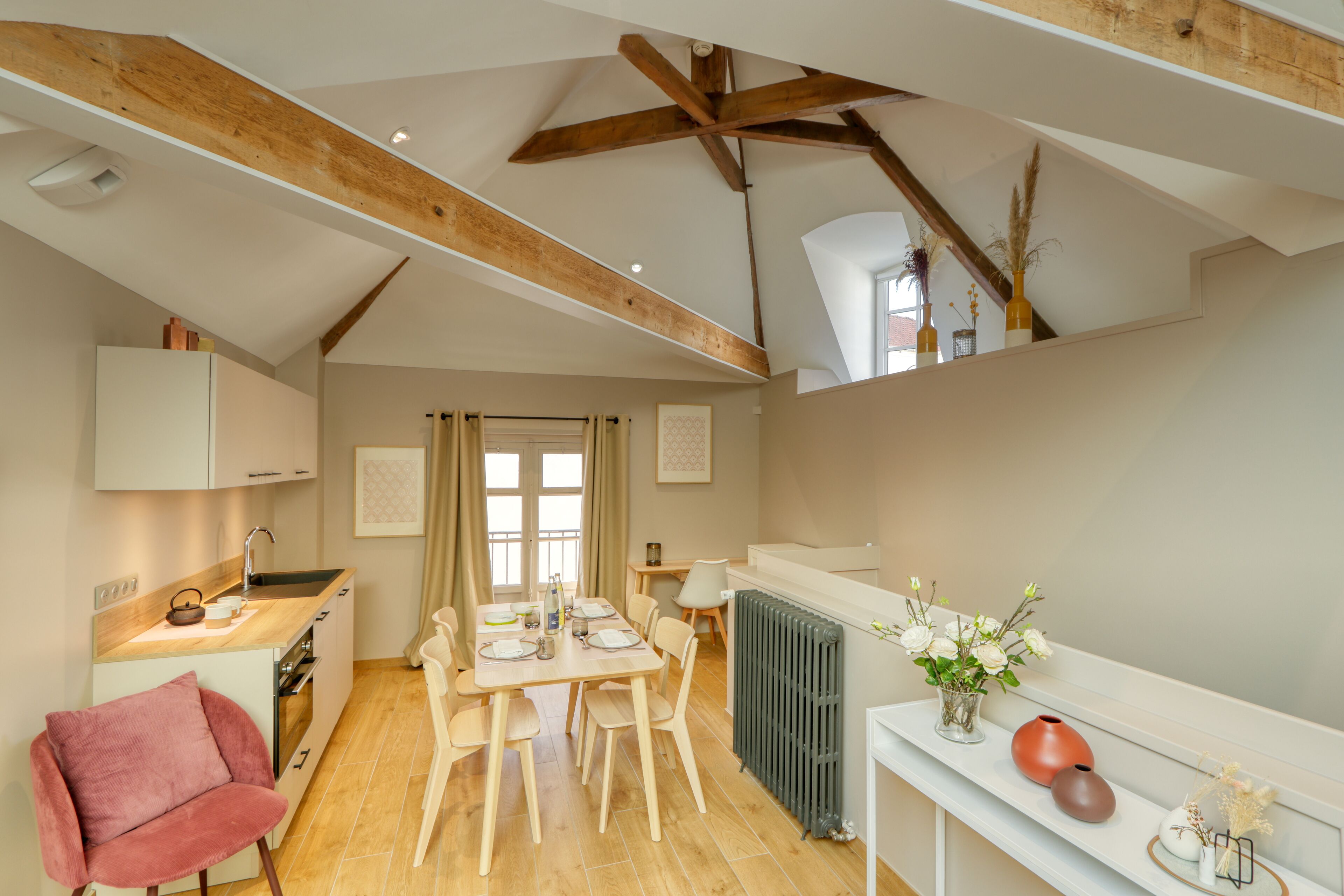 A spacious kitchen with wooden beams, a dining area with a table set for four, and a cozy seating area with a red chair.