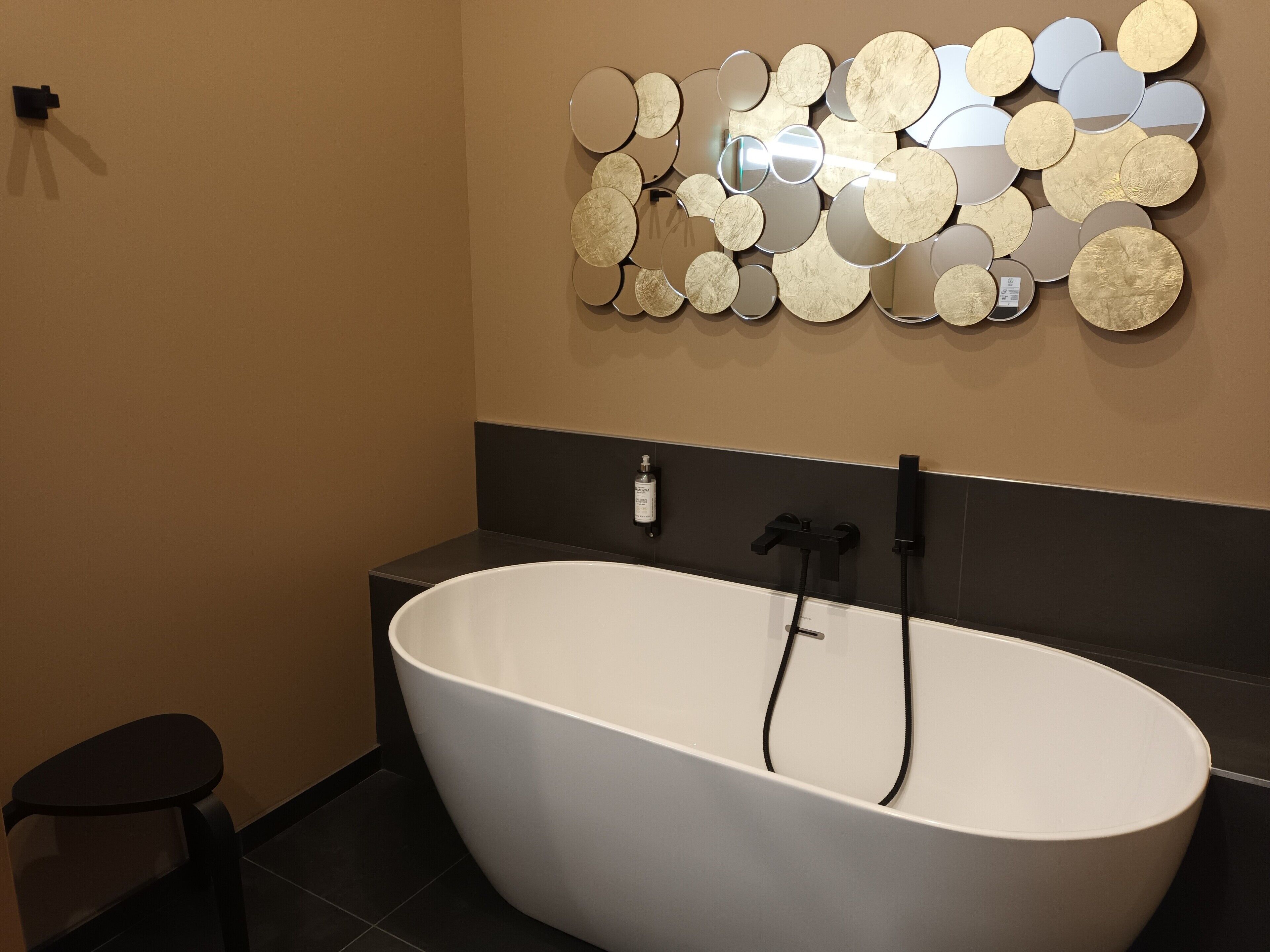 A modern bathroom with a freestanding bathtub, a wall-mounted mirror with circular patterns, and a small black stool.