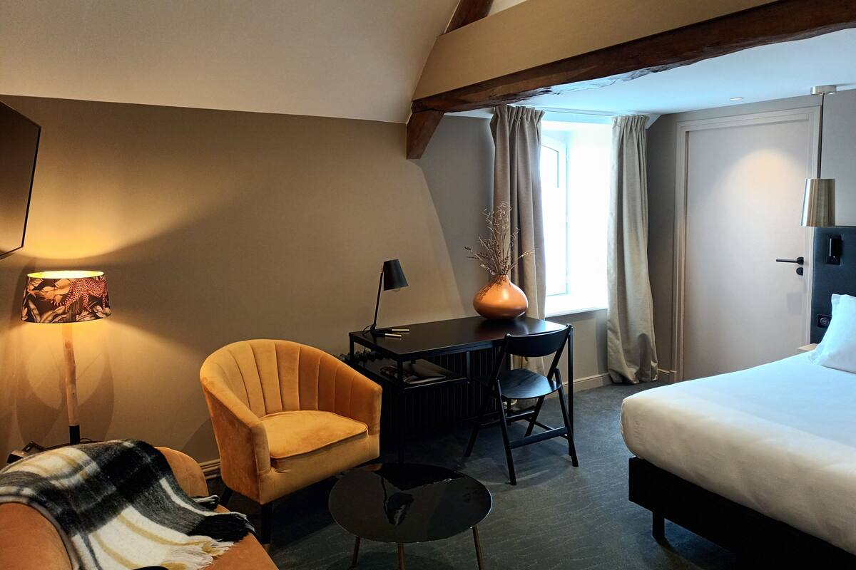 Habitación de hotel con cama, escritorio, silla, mesita, lámpara y un jarrón con flores.