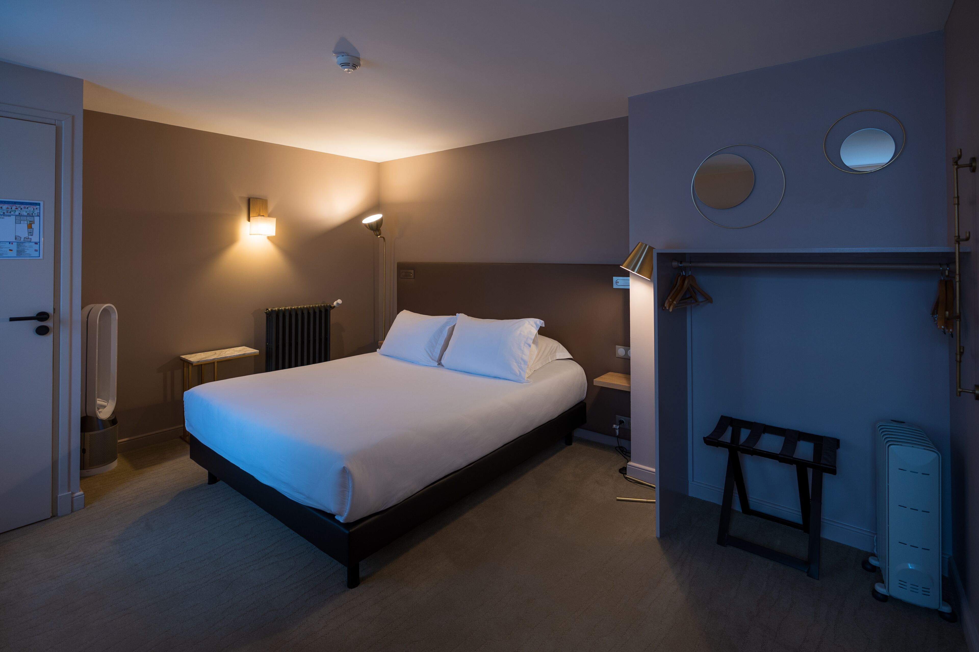 A hotel room with a bed, bedside lamps, a radiator, a coat rack, and a small table.