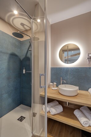 Un baño moderno con ducha envuelta en vidrio, un espejo redondo y un mueble de tocador de madera con lavamanos blanco.