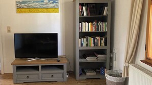 En stue med et fladskærms-TV på et træstel, en reol fyldt med bøger, et billede på væggen og et vindue med gardiner.