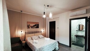A bedroom with a bed, bedside lamps, a nightstand, a chair, a painting, and a bathroom with a sink and mirror. - Brand new apartment adjacent to the famous Vitelli bar from the film IL PADRINO. (Savoca)