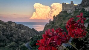 A coastal landscape with a castle-like structure on a hill, a winding road, and red flowers in the foreground. - Brand new apartment adjacent to the famous Vitelli bar from the film IL PADRINO. (Savoca)