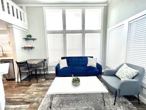 A living room with a blue sofa, a grey sofa, a white coffee table, and a grey rug. There is a small table with chairs and a plant on the coffee table.