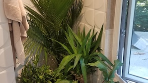 A corner of a room with a window, a white tiled wall, and a plant shelf with various plants.