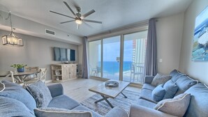 A modern living room with a large gray sectional sofa, a wooden coffee table, and a ceiling fan. There is a balcony with a view of the ocean and a dining area with a table and chairs.