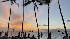 A beachside restaurant with outdoor seating, palm trees, and a view of the ocean at sunset. - Luxury Zen Polynesian Penthouse (Waikoloa Resort)