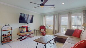 A living room with a TV, a sofa, a rocking chair, a wooden table, and a balcony with curtains. - Beach Therapy at Maravilla by My Galveston Getaway (Galveston)