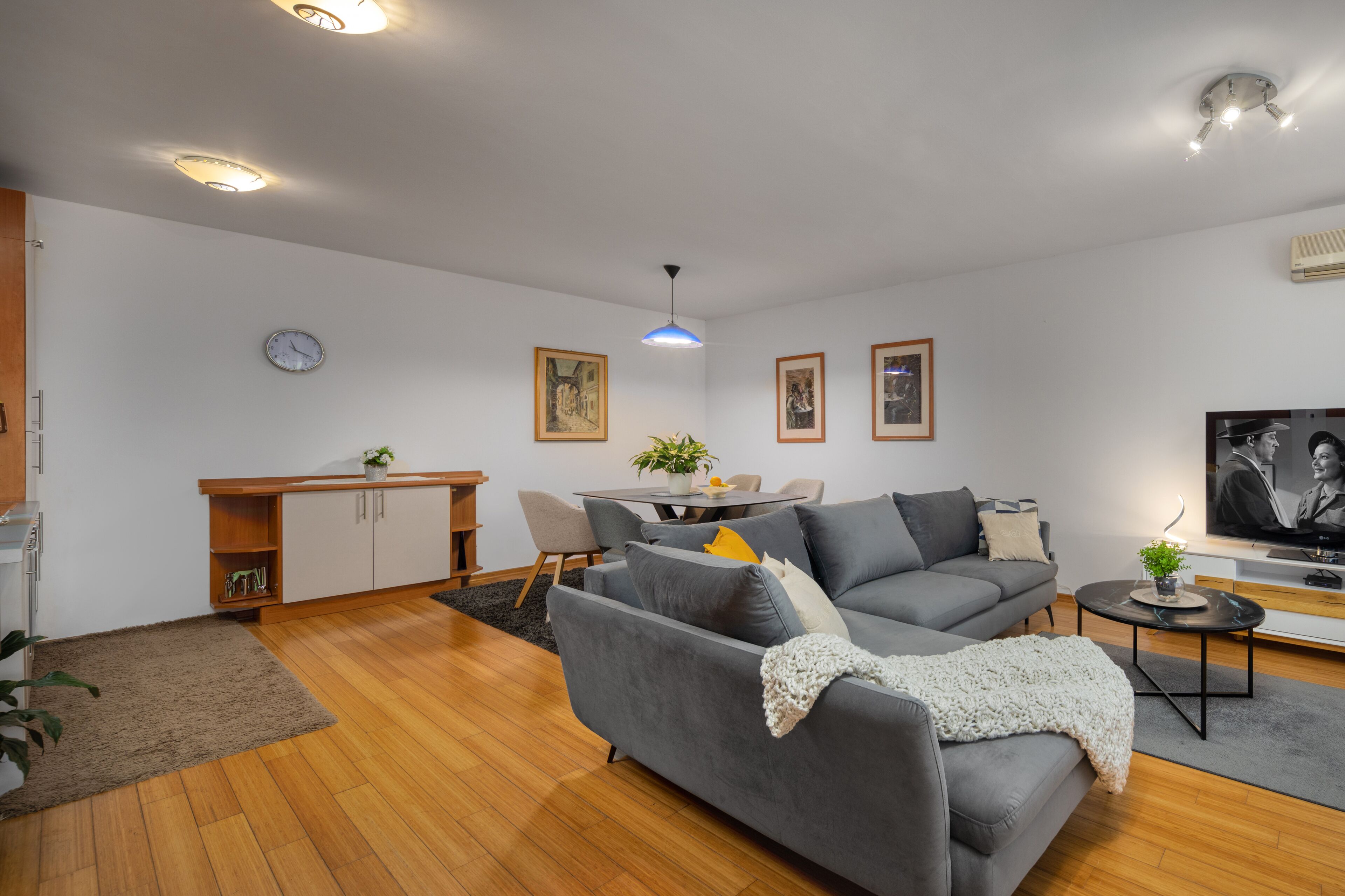Un salon moderne avec un canapé gris, un plancher en bois, une table à manger avec des chaises, un petit coin cuisine et un téléviseur à écran plat.