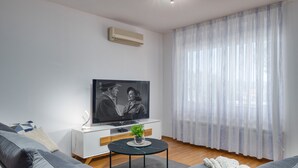 En moderne stue med sofa, sofabord, fjernsyn og et stort vindue med lette gardiner.