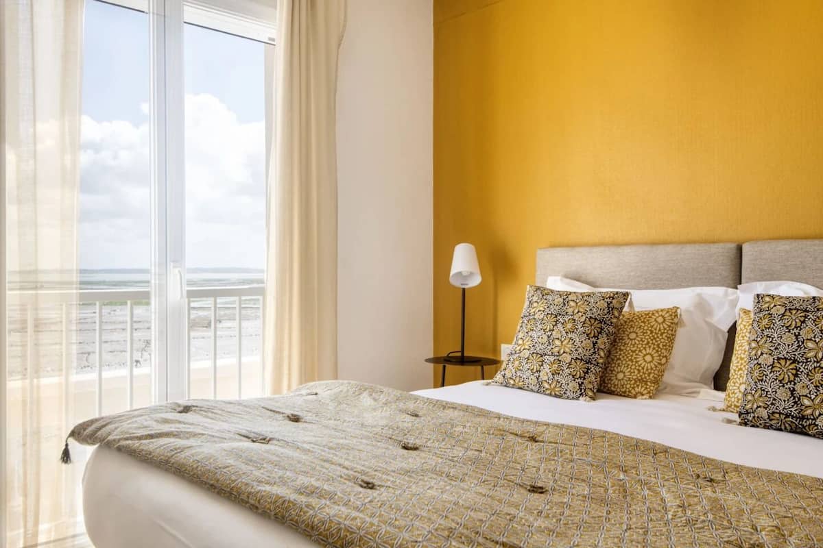 A hotel room with a large bed, a view of the beach, and a yellow wall.