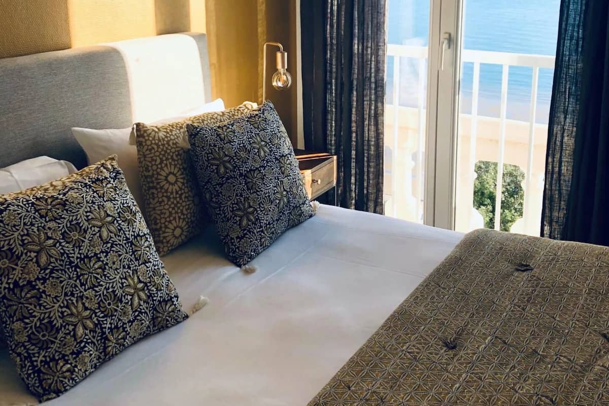 A hotel room with a bed, pillows, a nightstand, and a view of the sea through a large window.