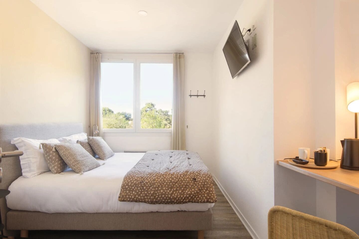 Une chambre d’hôtel comprenant un lit, un bureau, une chaise, un téléviseur fixé au mur et une fenêtre avec des rideaux.