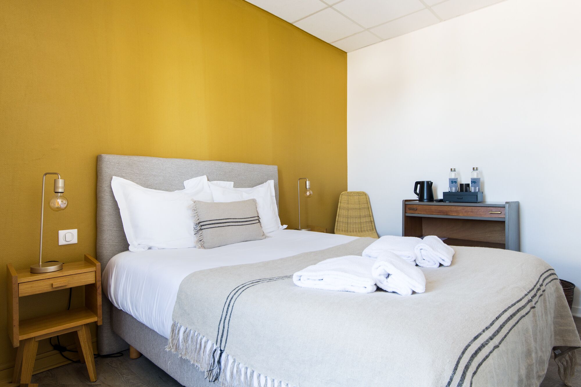 A neatly made bed with white linens and a beige throw blanket, a bedside table with a lamp, a chair, and a small desk with a coffee maker and water bottles.