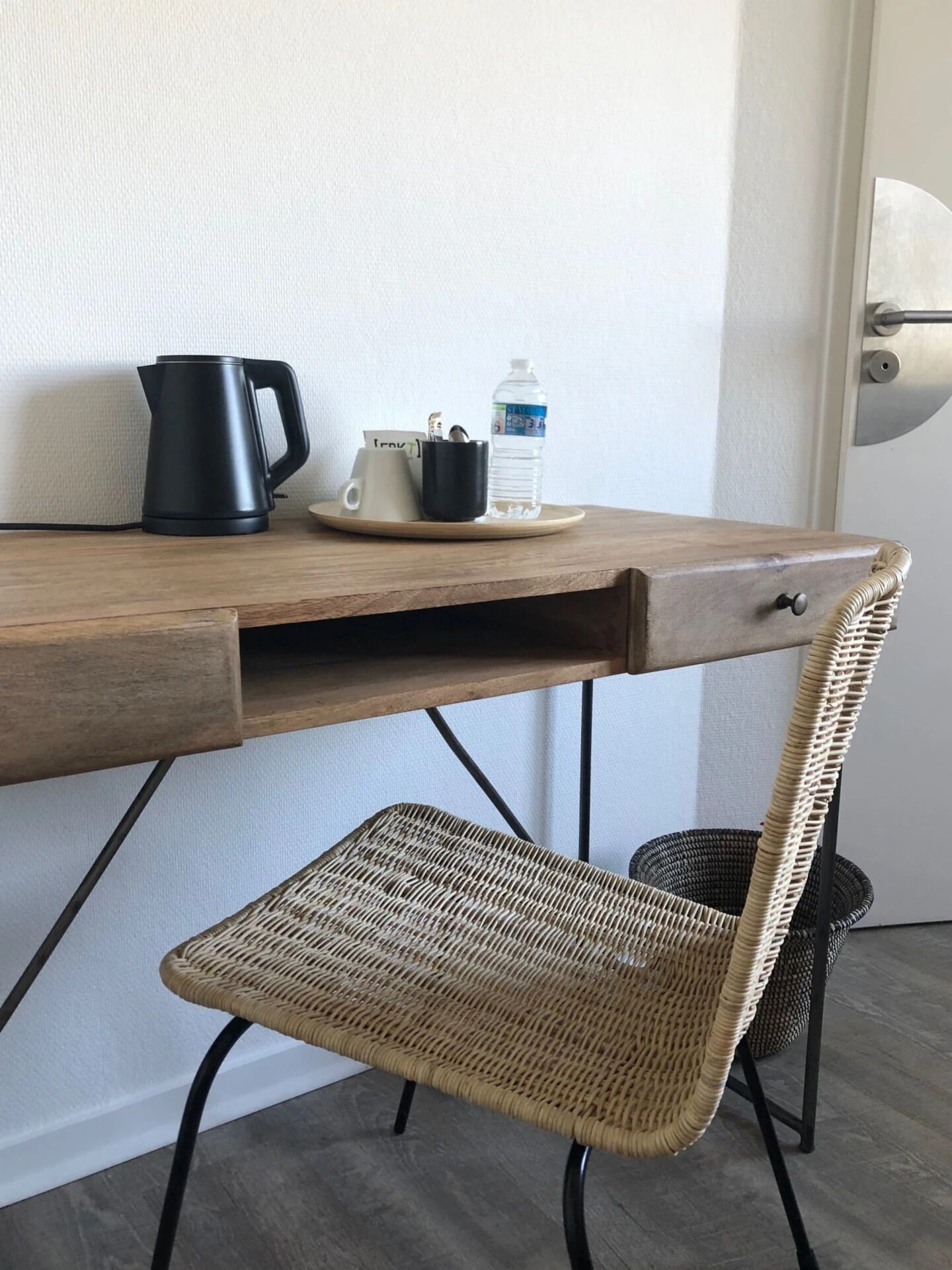 A wooden desk with a black kettle, a water bottle, and a mug. Below the desk is a wicker chair.